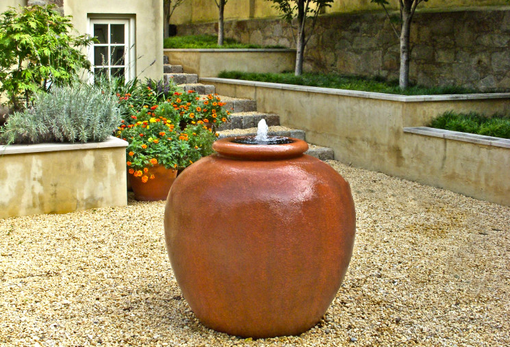A rich orange vessel fountain is located in bed of pea gravel